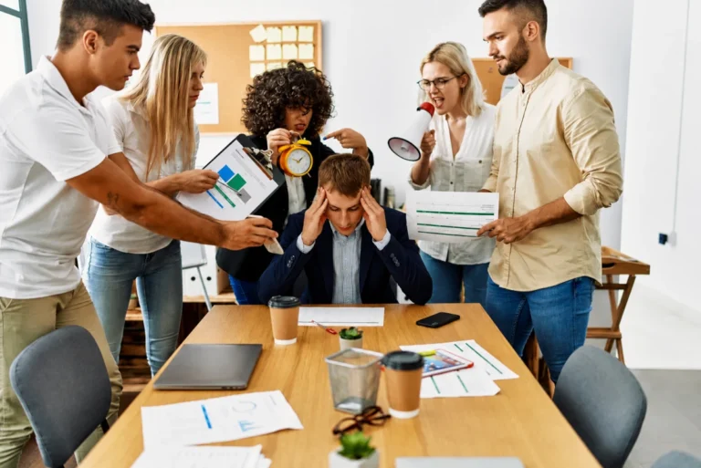 Empleado visiblemente agotado mientras varios compañeros le presionan con tareas y demandas en una oficina, representando el estrés laboral y el burnout colectivo en equipos de trabajo.